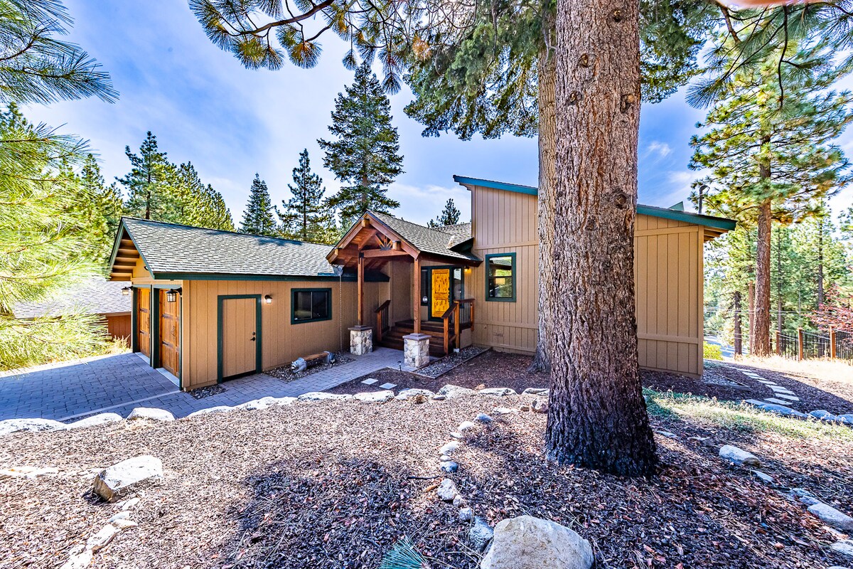 The exterior of the newly renovated mountain home showcases a welcoming entrance under a rustic wooden overhang. Surrounded by mature pine trees, the home is set on a landscaped lot with a stone pathway leading to it, offering a serene mountain retreat.
