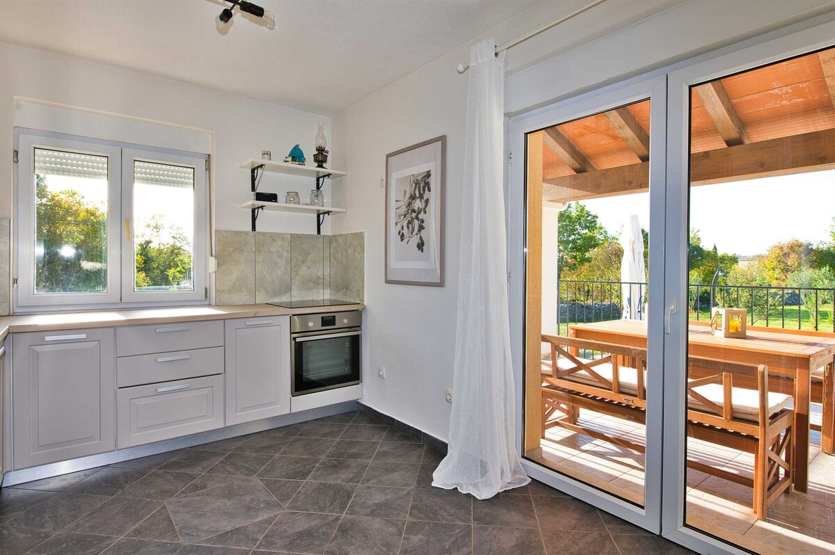 A well-equipped kitchen features modern cabinetry and appliances, including an oven. Large windows provide views of the outdoor dining area, which is furnished with a wooden table and chairs under a shaded terrace.