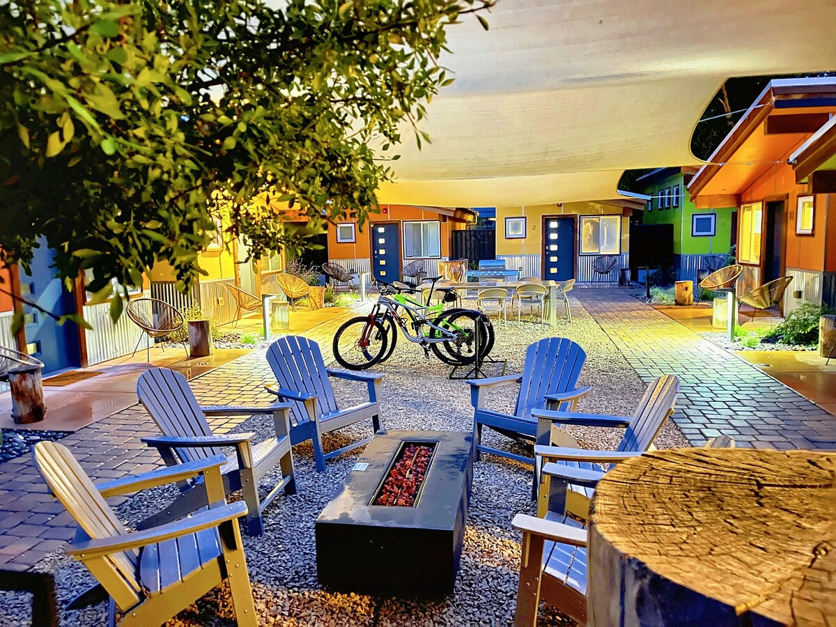 An outdoor gathering area features multiple Adirondack chairs arranged around a circular fire pit. Two bicycles are parked nearby, and pathways among greenery lead to colorful studio units. Soft lighting enhances the welcoming atmosphere of the shared courtyard.