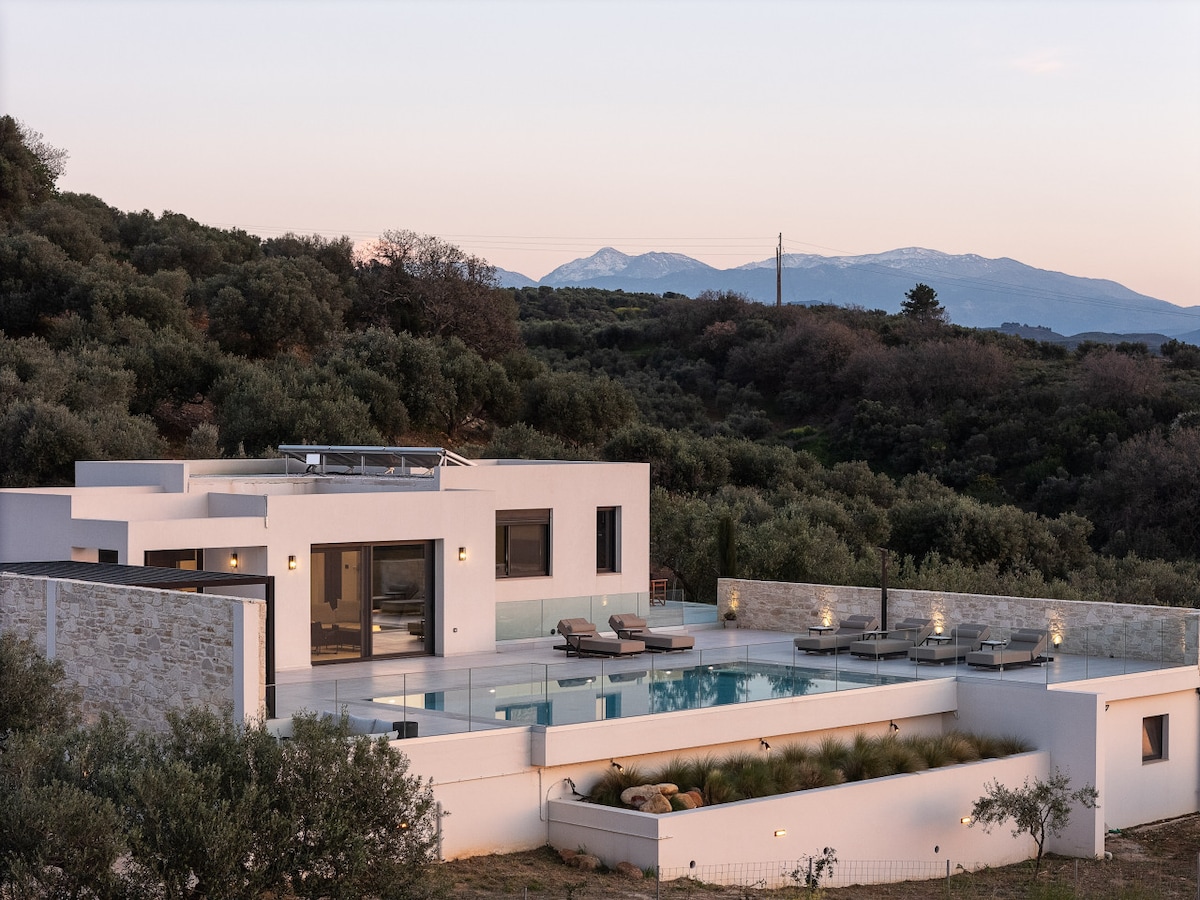 The villa is positioned on a hillside, featuring a private heated pool surrounded by sunbeds. Olive trees dot the landscape, with distant mountains visible beyond. The modern architecture of the villa complements the serene natural setting, emphasizing a secluded atmosphere.