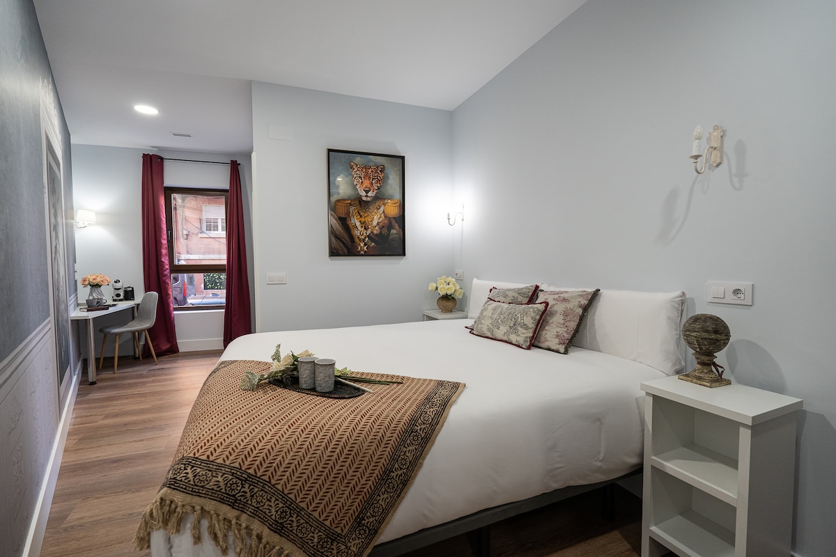 A spacious room features a comfortable bed adorned with a patterned throw. A small table is positioned nearby, and a window dressed with burgundy curtains allows natural light. Wall art and soft lighting fixtures enhance the overall ambiance while wooden flooring completes the inviting setting.