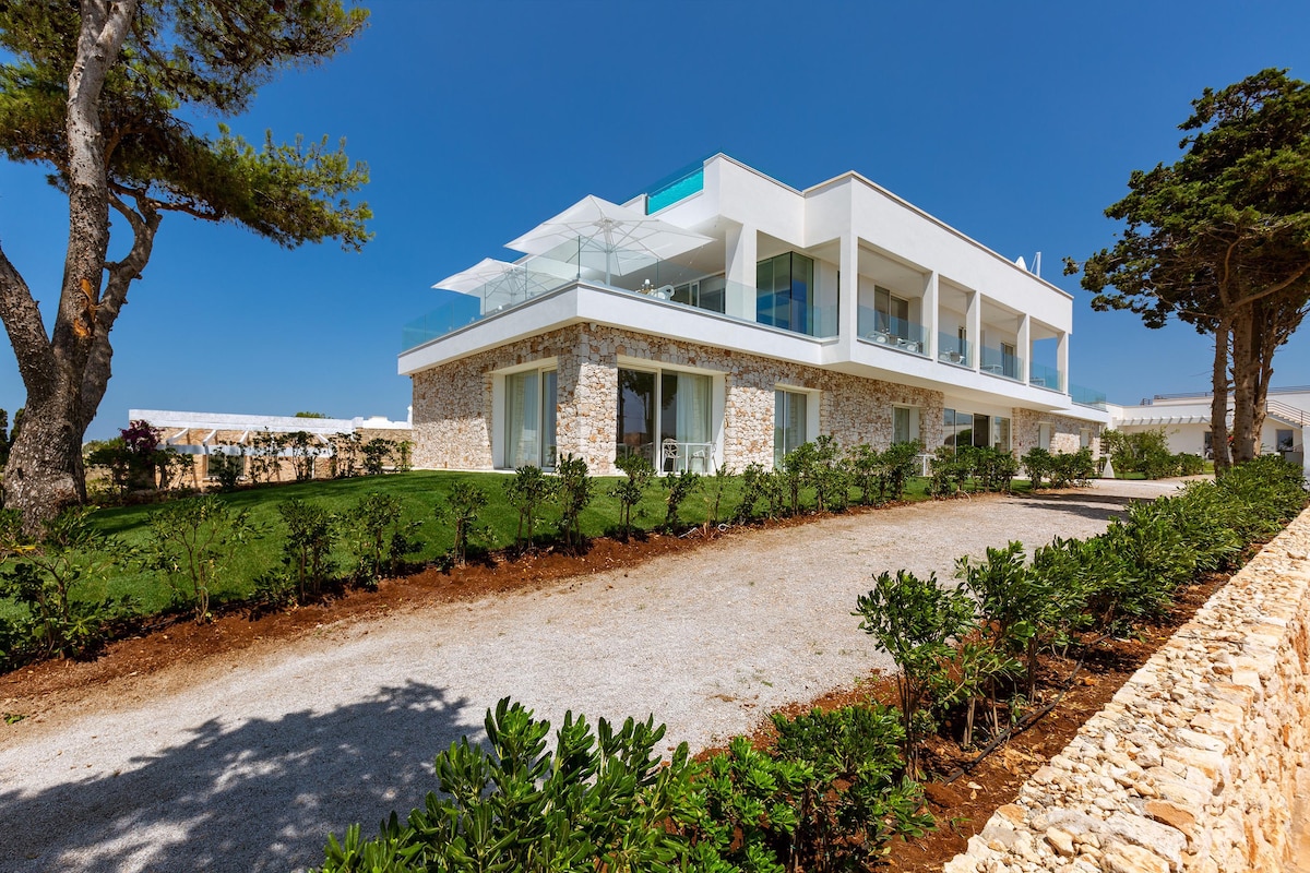 A modern, multi-level building is surrounded by well-manicured greenery and features a spacious balcony area. Large glass windows reflect the clear blue sky, while a gravel pathway leads up to the entrance. Sunlight enhances the contemporary architectural lines of the property.