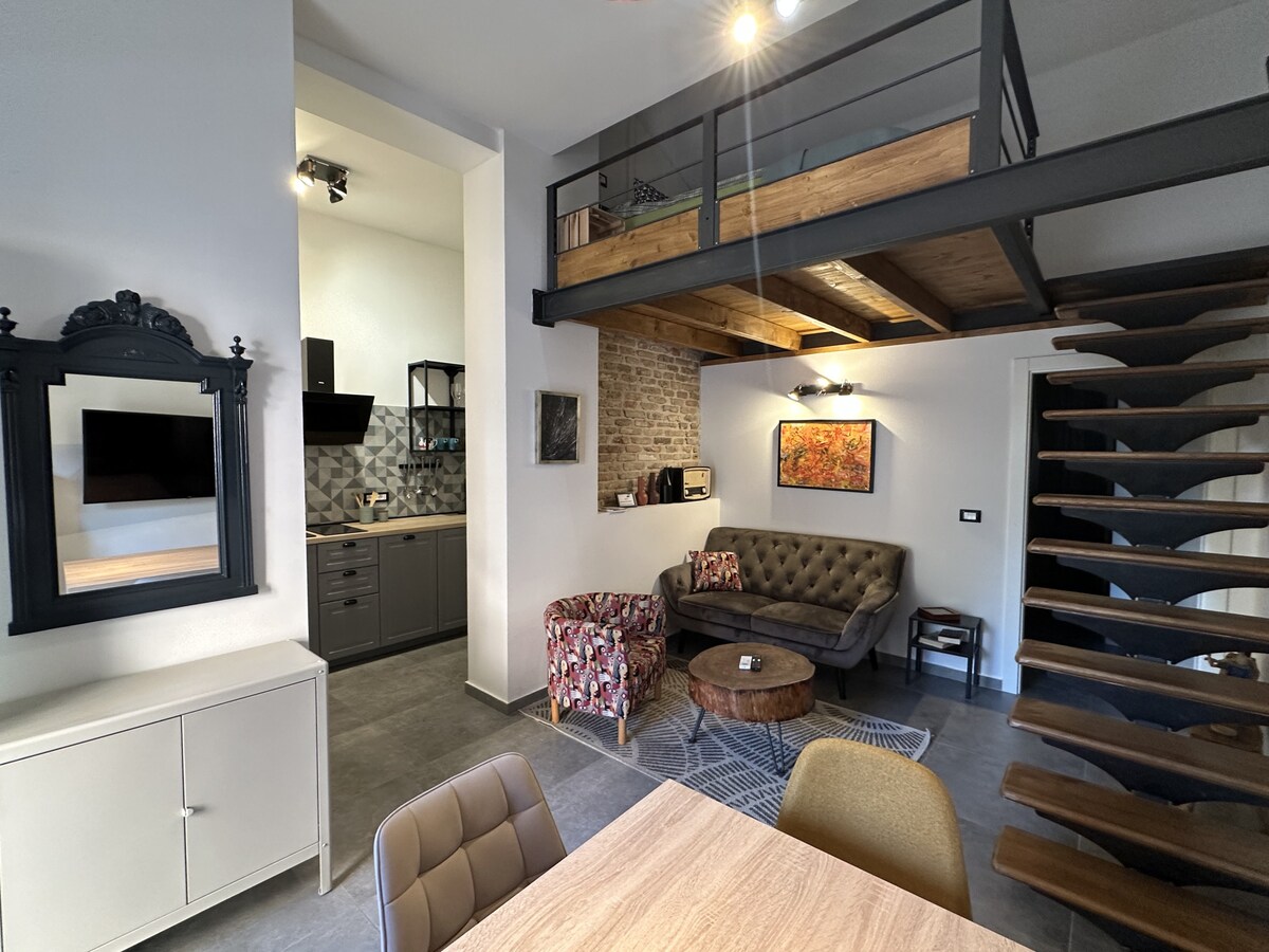 An open living area features a comfortable seating arrangement beside a coffee table and a staircase leading to a lofted space. The kitchen is visible, showcasing modern appliances and cabinetry. A framed artwork adds a decorative touch to the brick wall.