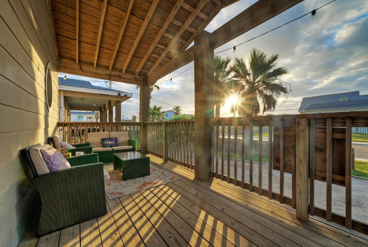 The outdoor deck features comfortable seating surrounded by wooden railings, with sunlight streaming through the slats. A warm glow is created by the soft illumination of string lights overhead, while palm trees can be seen in the background under a partially cloudy sky.