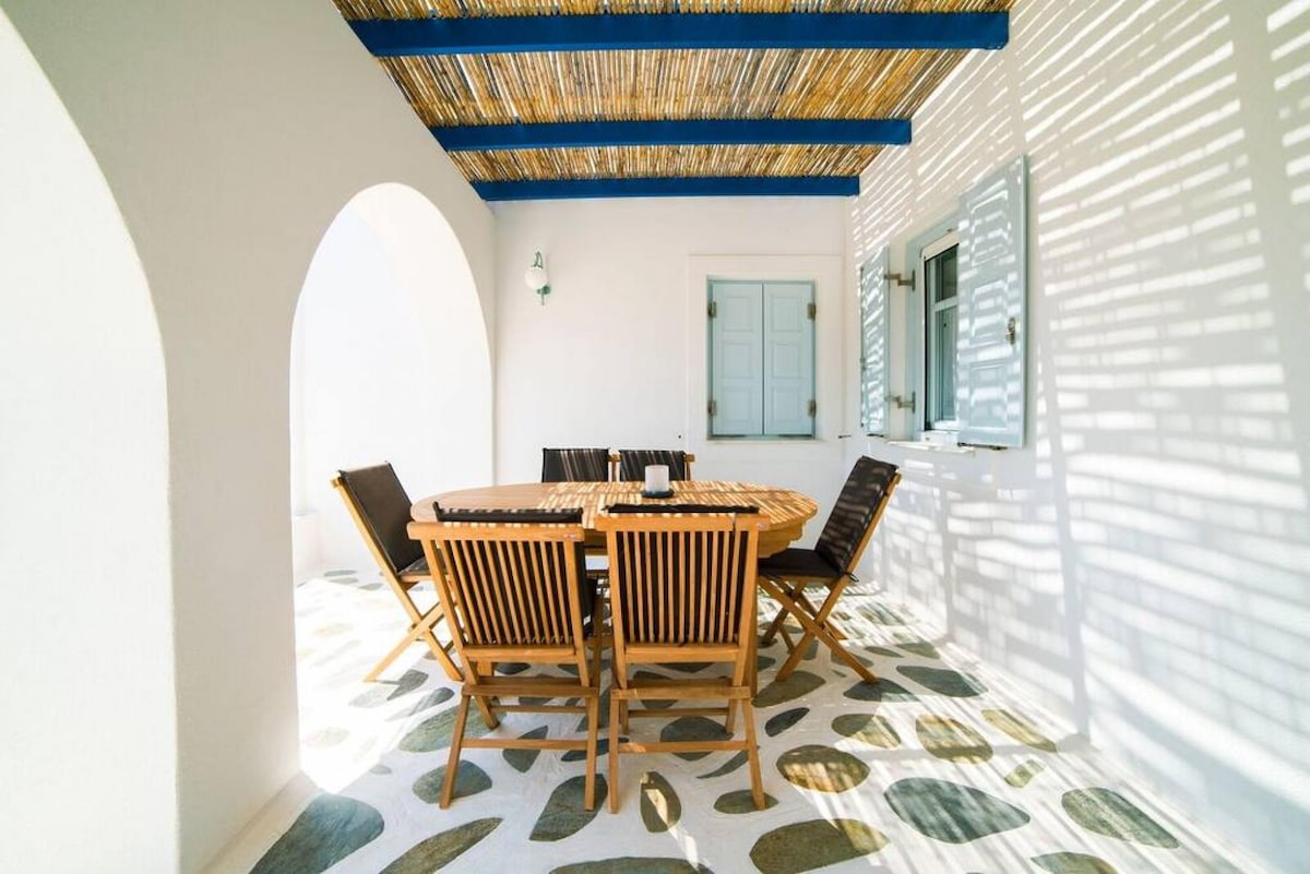 An outdoor dining terrace is presented with a large round wooden table surrounded by six wooden chairs. A thatched roof provides partial shade, while sunlight casts patterned shadows on the surface. The area features white walls and a light blue door, enhancing the space.