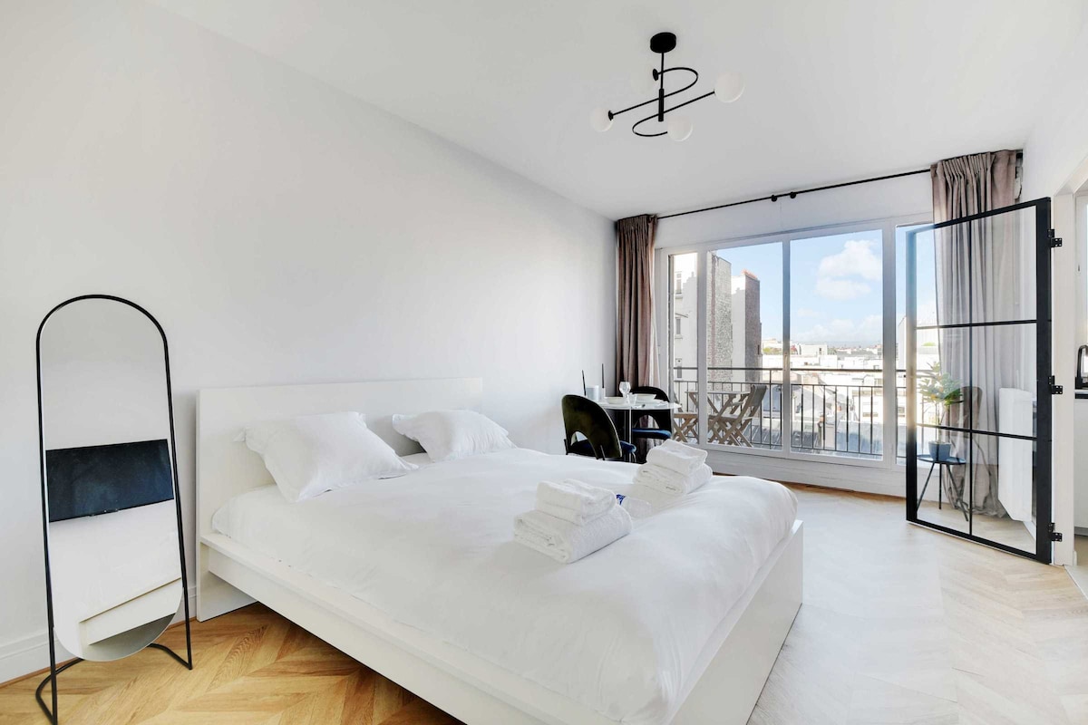 A bright studio space features a spacious double bed dressed in white linens. Near the bed, a large window offers natural light, complemented by elegant curtains. A stylish mirror and a small dining space with a chair are thoughtfully arranged, creating a serene environment.