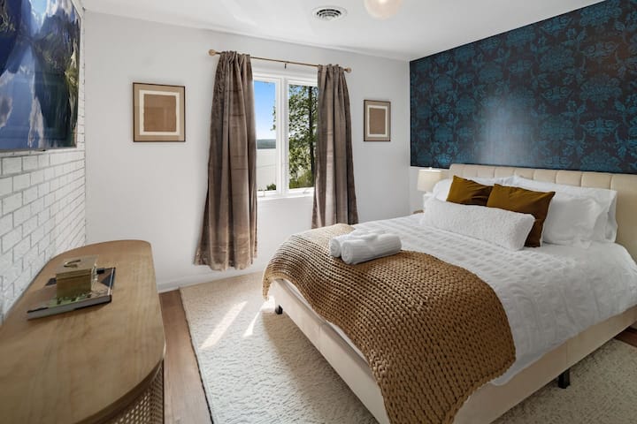 Un dormitorio luminoso y acogedor con una cama cómoda adornada con almohadas blancas y mostaza, un tiro texturizado, una elegante pared decorativa azul y un escritorio de madera, todo iluminado por luz natural desde una ventana que ofrece una vista al aire libre.