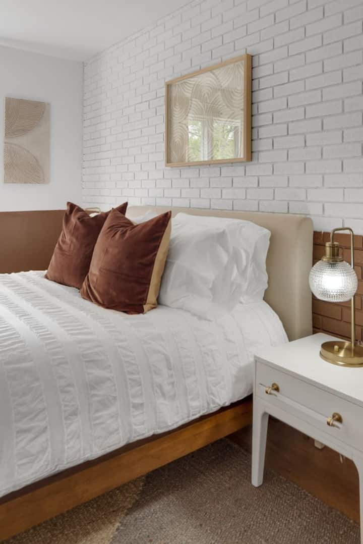 Un dormitorio acogedor con una cama lujosa, ropa de cama blanca, almohadas decorativas marrones, una pared de estilo ladrillo, un arte de pared abstracto, una mesita de noche clásica con una lámpara dorada, todo lo que contribuye a un ambiente cálido y acogedor.