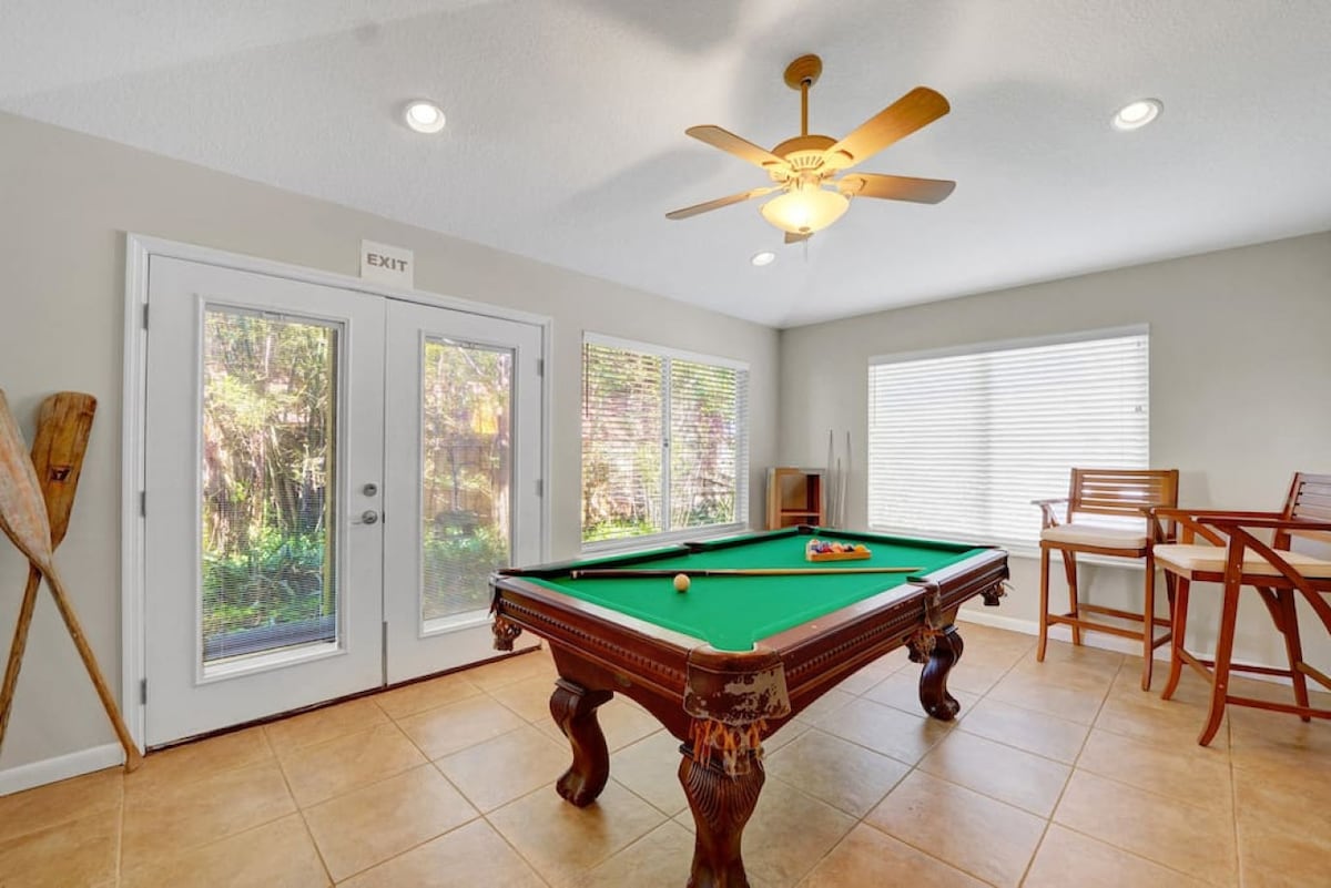 A game room is shown, featuring a billiard table with a cue ball and rack positioned on top. Natural light streams through large windows onto tile flooring. Two high-backed chairs are situated near a small table, creating a relaxed seating area. A ceiling fan is visible above.