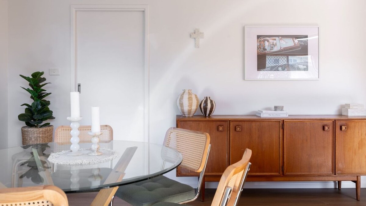 A dining area features a round glass table surrounded by four wooden chairs. A wooden sideboard displays decorative vases and a framed artwork. A potted plant enhances the corner, while natural light streams in, highlighting the clean, contemporary design.