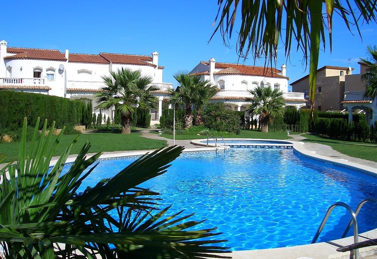 A shared outdoor area features a large swimming pool surrounded by lush green lawn and palm trees. Nearby, buildings with Mediterranean architecture are visible, providing a sense of tranquility and relaxation.