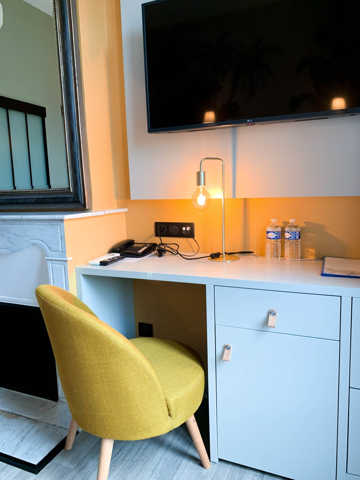 A workspace is presented with a light-colored desk featuring a sleek modern design. A yellow chair offers a pop of color. A flat-screen television is mounted above, while a lamp provides soft illumination. Two bottles of water are neatly placed on the desk.
