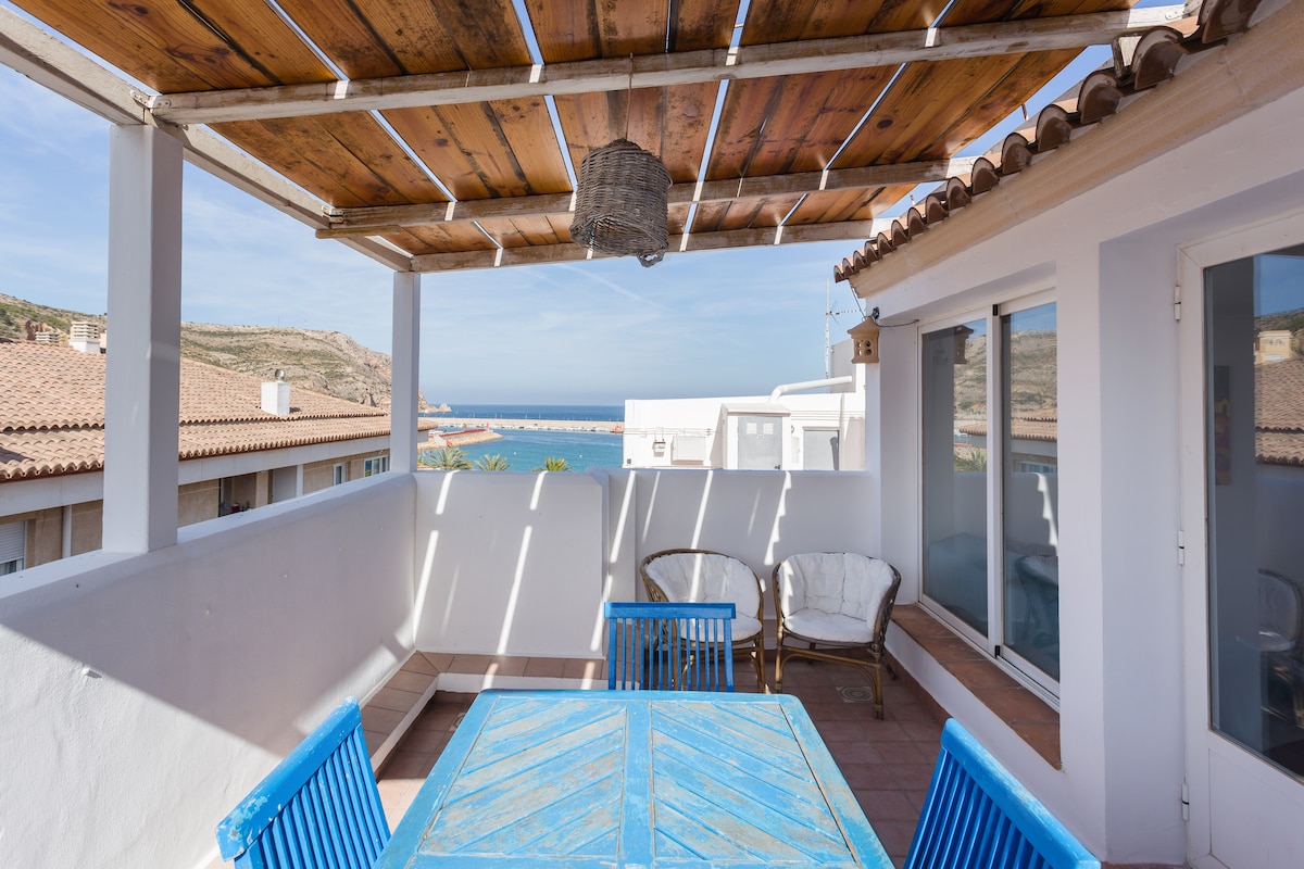 A private covered terrace is depicted, featuring a blue table and matching chairs. Sunlight highlights the space, with views of the sea visible in the background. Overhead, wooden beams support a slatted roof, enhancing the open and airy feel of the area.