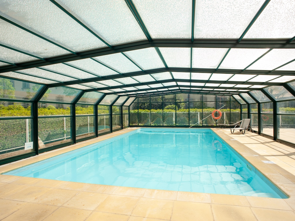 A covered outdoor pool area is visible, featuring clear walls and ceiling. The water is calm and inviting, reflecting light from above. Lounge chairs are positioned nearby on the tiled deck, offering a place to relax while enjoying the warm environment.