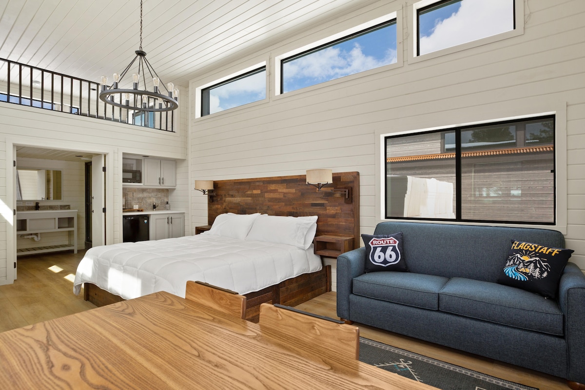 A spacious bedroom features a king-sized bed with crisp white linens and a wood-paneled accent wall. A comfortable blue couch is positioned nearby, and large windows allow natural light to fill the room, showcasing the wood flooring and a dining area in the foreground.