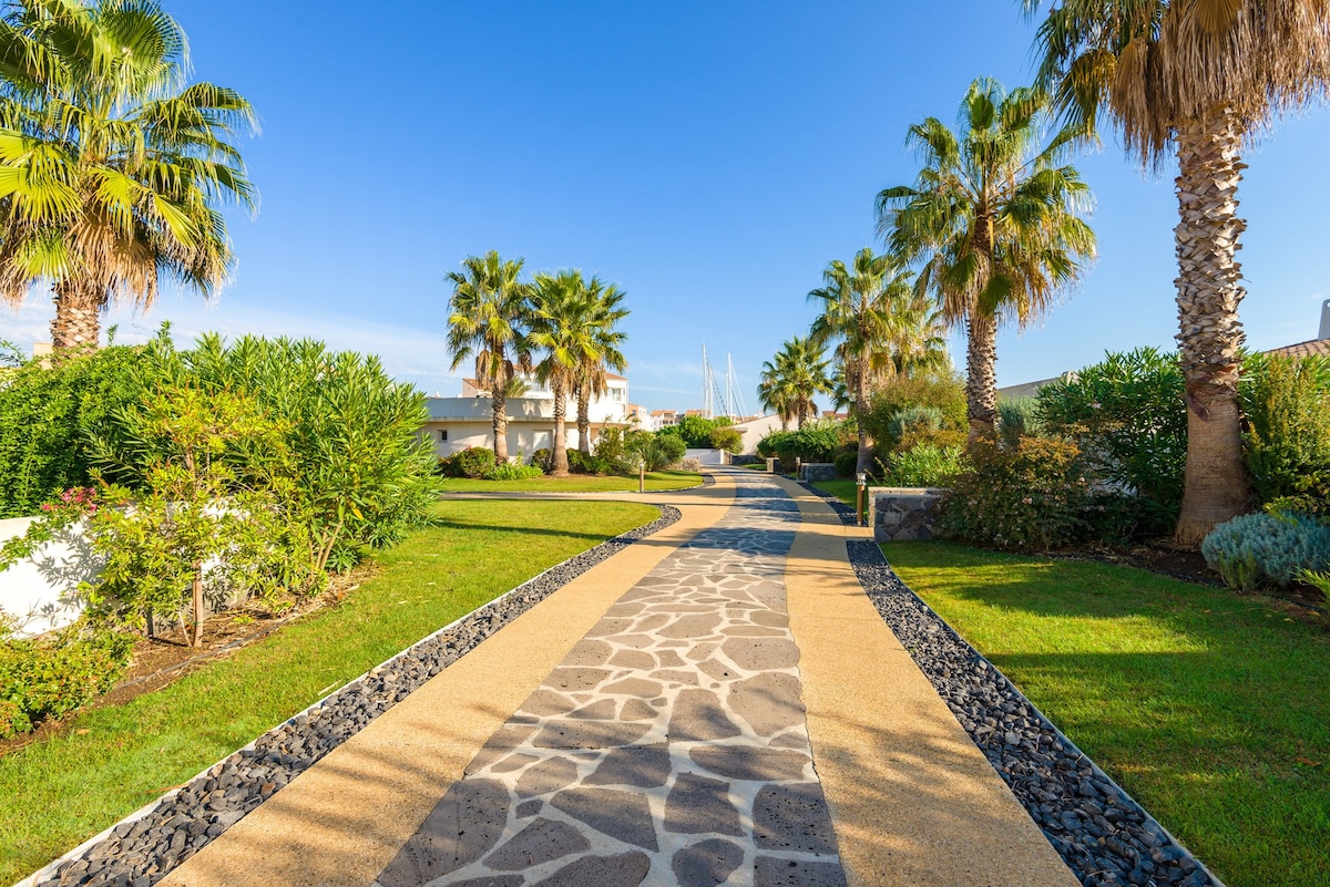 A meandering pathway lined with palm trees leads through lush greenery, creating a welcoming atmosphere. The path is paved with stone and surrounded by vibrant plants, inviting guests to explore the area. In the background, boats are visible near the marina.