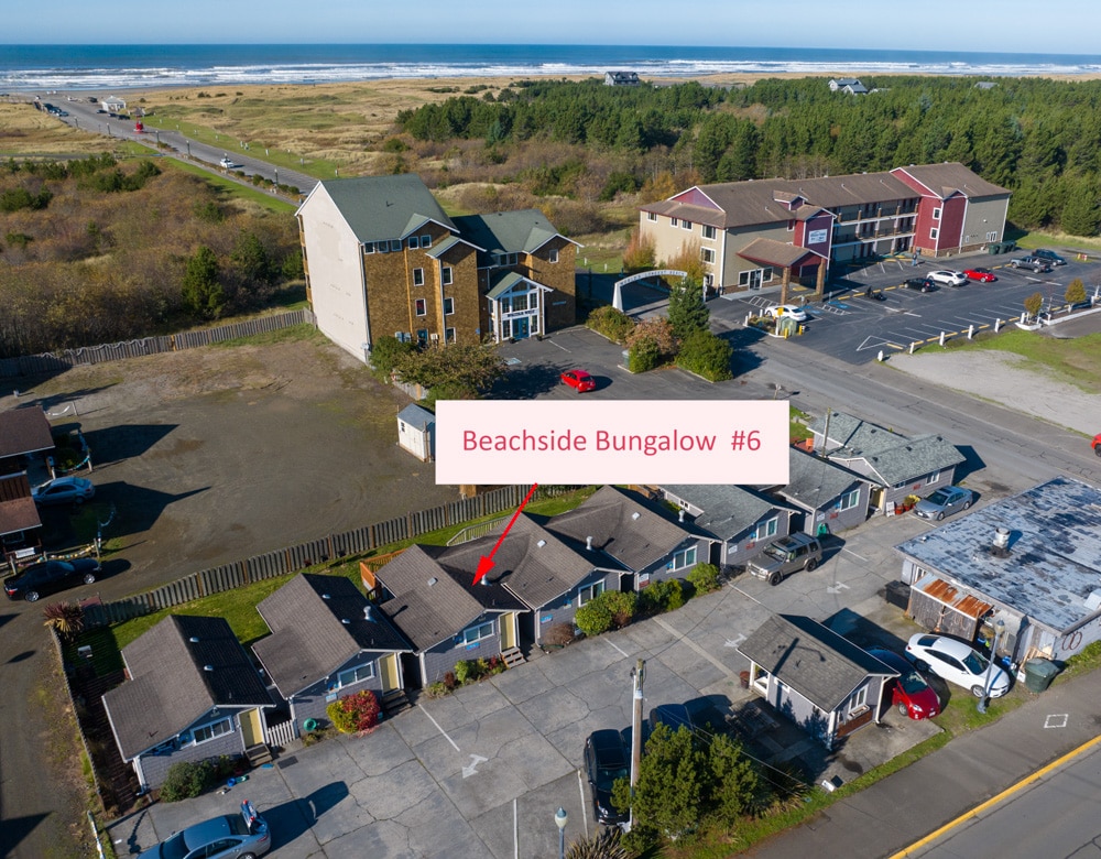 An aerial view captures Beachside Bungalow #6, nestled among a cluster of charming cabins. The surrounding greenery and paved area are visible, with the beach and ocean in the distance, illustrating the proximity to coastal experiences.