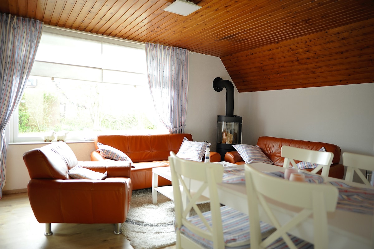 A living room features cozy orange leather sofas positioned around a white coffee table. A dining table with white chairs is visible, and a large window with light curtains allows natural light to fill the space. A modern wood-burning stove adds warmth to the room.
