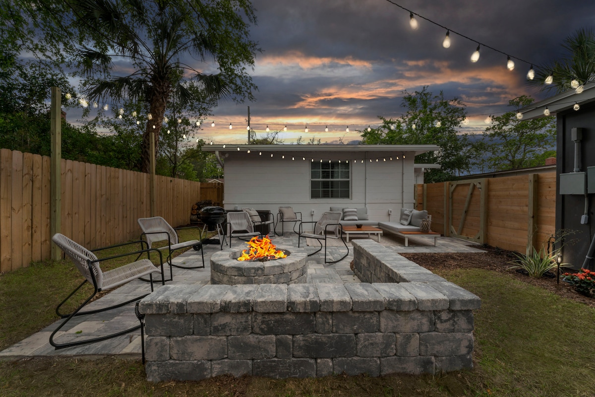 A cozy outdoor space features a circular stone fire pit surrounded by several comfortable chairs. Soft lighting hangs above, creating a warm ambiance. A wooden fence provides privacy, while a small patio area with additional seating is visible in the background, set against a tranquil sunset sky.