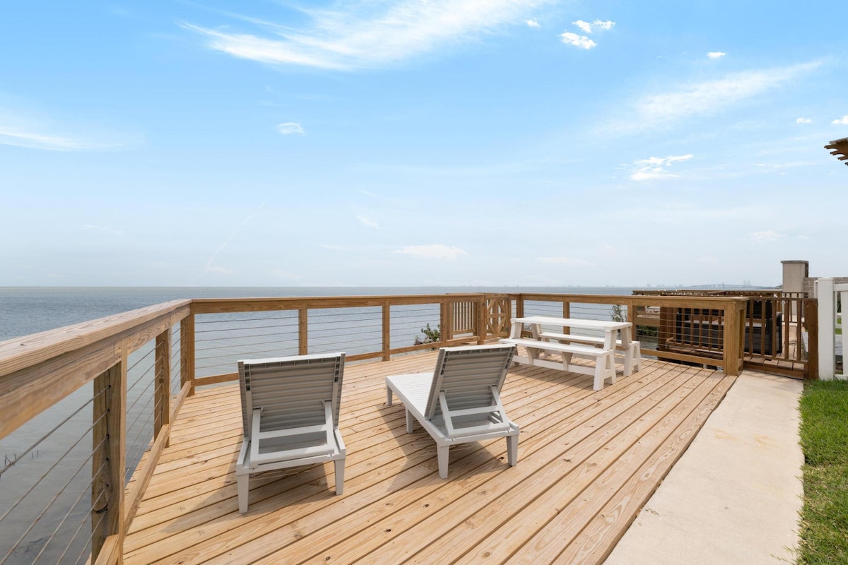 A wooden deck features two lounge chairs and a picnic table, providing an open area with unobstructed views of the water. The soft waves can be seen in the distance, complemented by a clear blue sky.