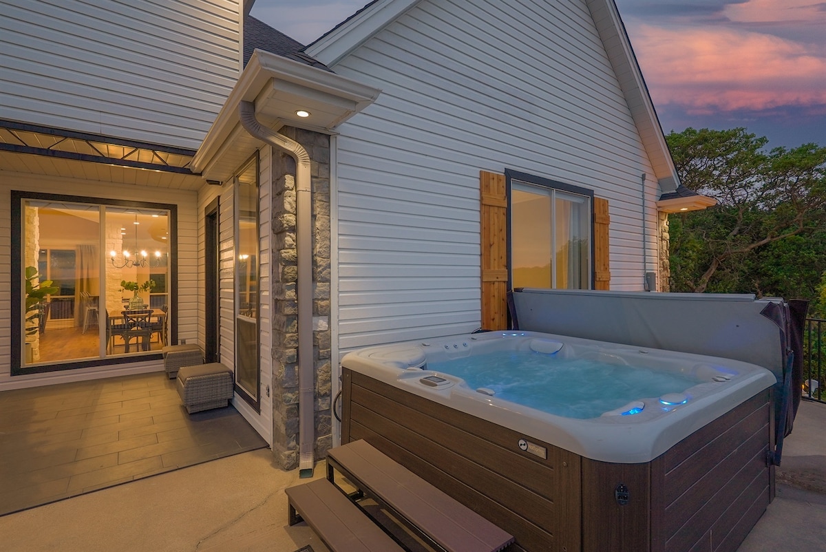 A private hot tub is positioned on a patio, surrounded by stone and wood accents. The serene waters of Possum Kingdom Lake can be seen in the distance, while large glass doors lead to an indoor dining area, enhancing the connection between indoor and outdoor spaces.