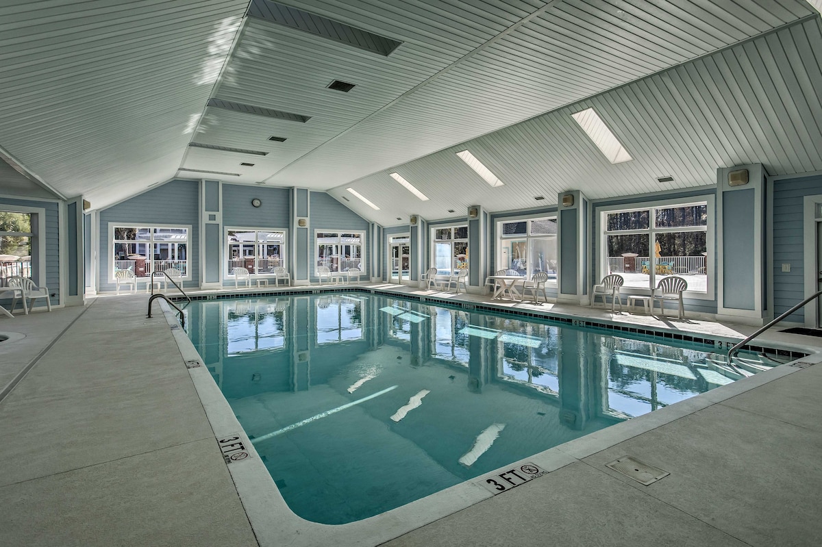 An indoor pool area features large windows allowing natural light to fill the space. The pool is surrounded by light-colored tile and complemented by several white lounge chairs, creating a bright and inviting atmosphere for relaxation.