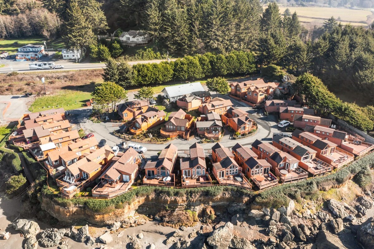 An aerial view captures a cluster of cabins nestled among lush greenery and rocky terrain. The layout forms a circular arrangement, surrounded by natural landscapes, with various buildings featuring red roofs and large windows. The nearby road and distant structures add context to the serene setting.