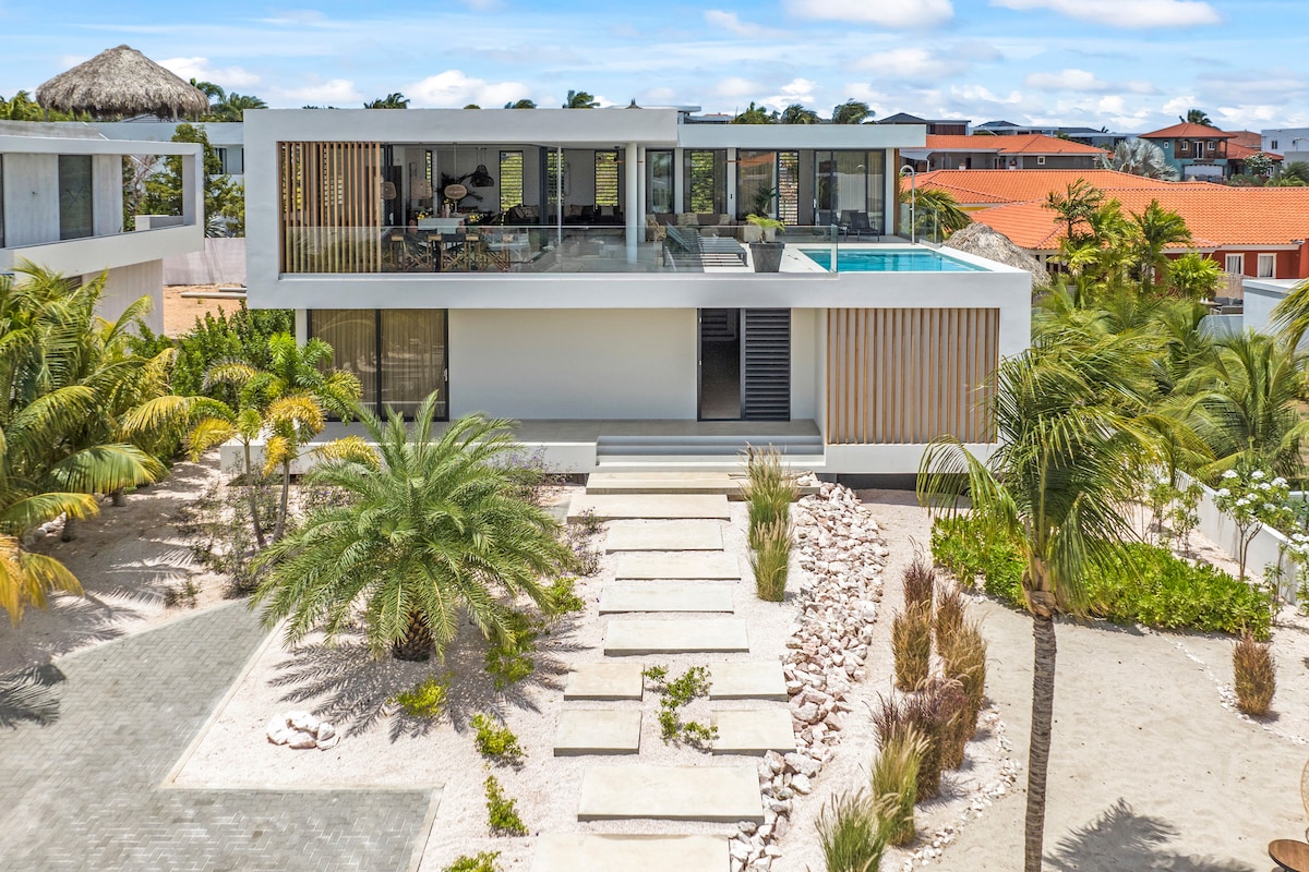 A modern villa is showcased from an aerial view, featuring a clean, white exterior with expansive glass windows. A private pool is visible on the upper level. Lush palm trees and decorative stones line the path leading to the villa, enhancing the tranquil outdoor space.