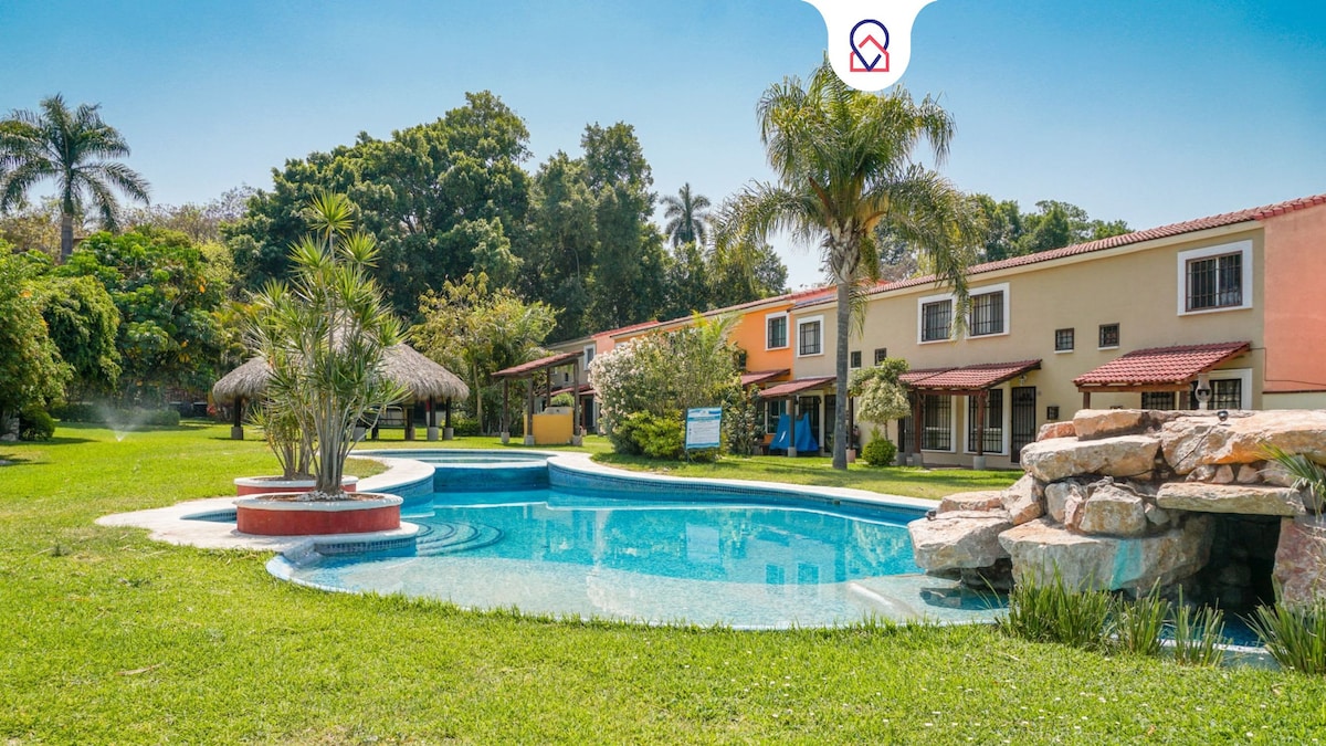 A serene outdoor area features a sparkling swimming pool surrounded by vibrant green grass and palm trees. A traditional palapa is visible nearby, offering shaded seating. The colorful residential buildings provide a picturesque backdrop, enhancing the peaceful ambiance of the setting.