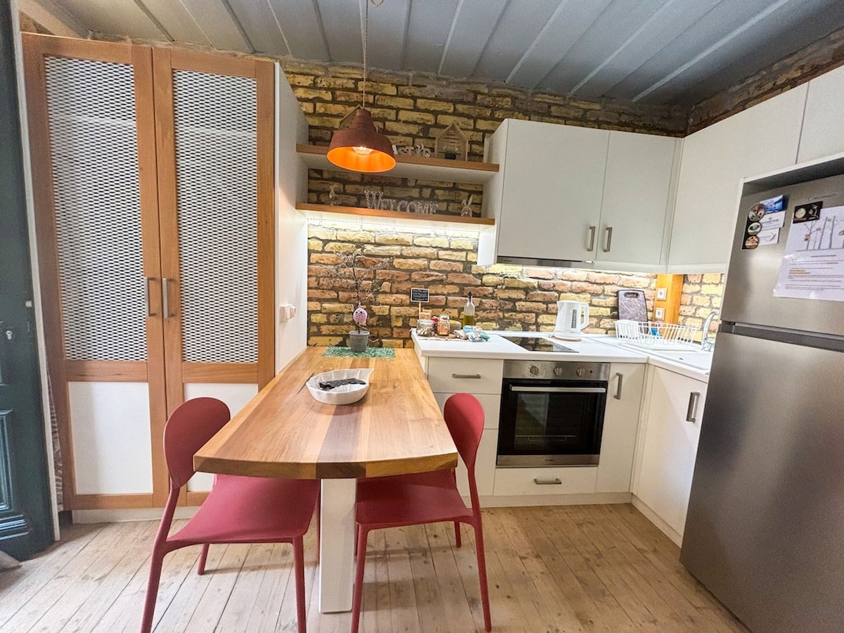 A modern kitchen features exposed brick walls, white cabinetry, and a wooden dining table with red chairs. Under-cabinet lighting highlights the workspace, which includes essential appliances such as an oven, refrigerator, and kettle, creating a practical setting for meal preparation.