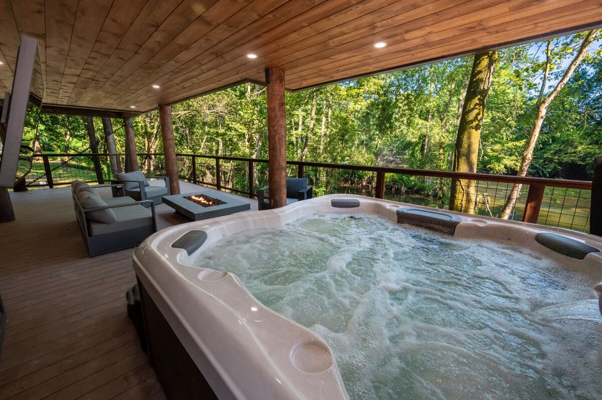 A hot tub filled with bubbling water is positioned on a covered deck, surrounded by wooden beams and expansive views of lush greenery. A cozy seating area with cushions and a fire pit table are visible, inviting relaxation in a tranquil outdoor setting.