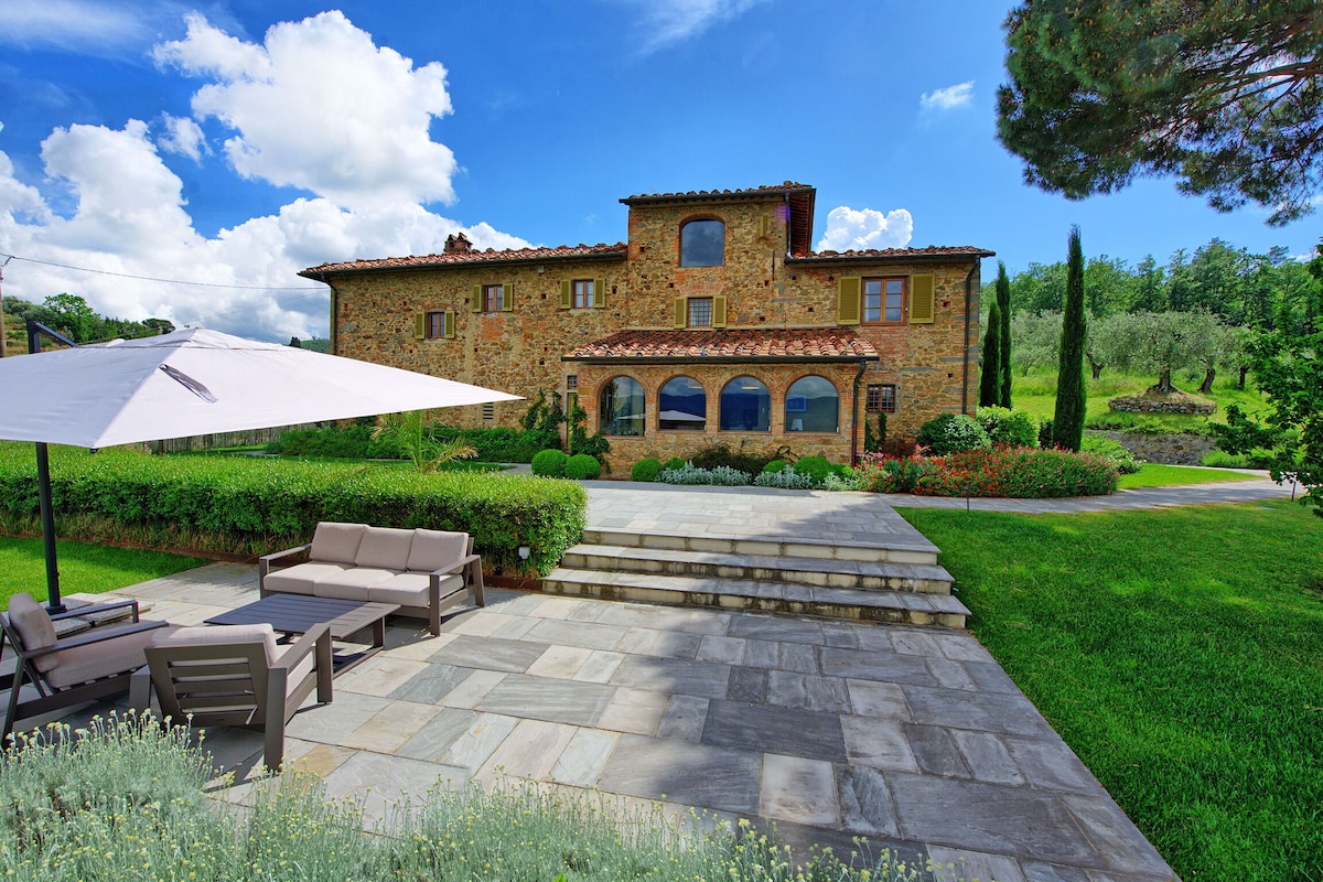 The villa's rustic stone façade is complemented by lush greenery and vibrant flowers in the garden. A shaded seating area with a large umbrella and comfortable sofas is positioned on the stone patio, inviting relaxation while enjoying the scenic surroundings.