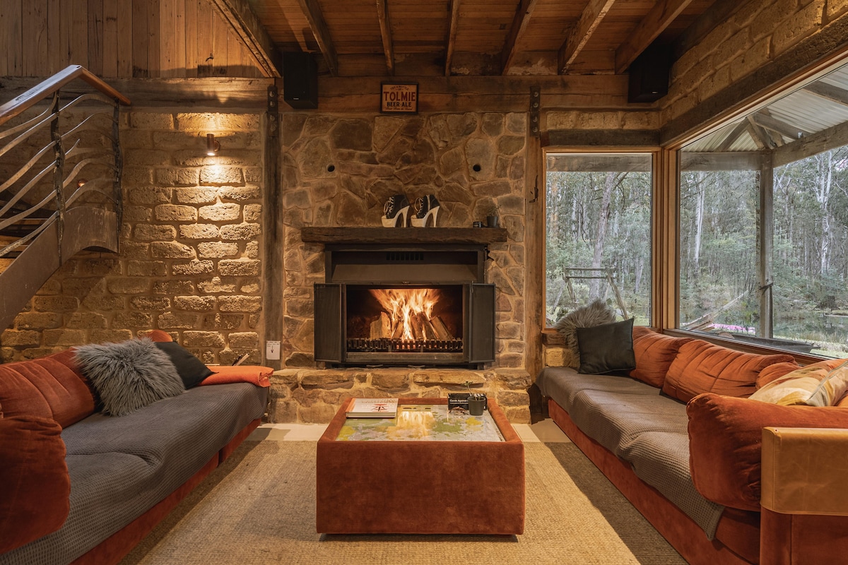 A warm living area features two comfortable couches positioned around a modern coffee table. The stone fireplace, with a vibrant fire, serves as a focal point. Large windows provide views of the surrounding bushland, enhancing the cozy atmosphere.