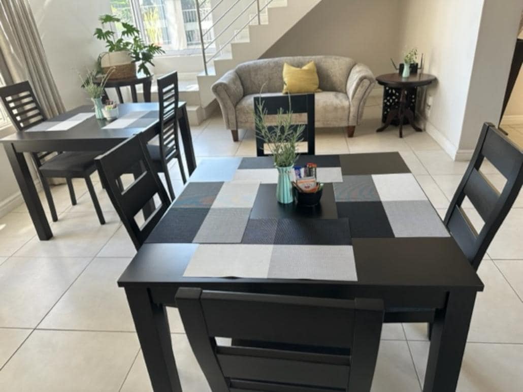 The dining area features two black wooden tables, each surrounded by matching chairs. A couch with neutral upholstery and a decorative yellow cushion is positioned nearby. Natural light fills the space through large windows, enhancing the overall ambiance.