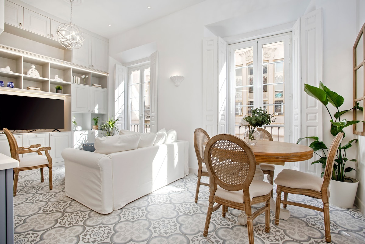 The living room features a light-colored sofa arranged near a bright window, enhancing the natural light. A round dining table with four chairs is positioned adjacent. Decorative plants are present, while a television and elegant shelving are situated in the background, providing functionality.