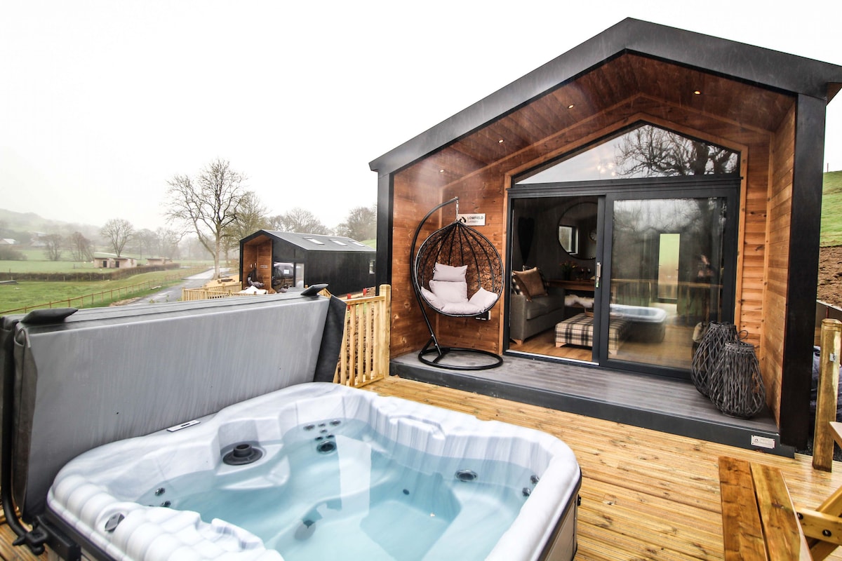 A wooden cabin exterior is complemented by a private hot tub on the deck, inviting relaxation. An ergonomic hanging chair, positioned nearby, offers a cozy spot to unwind. Large glass doors provide a view into the interior space, surrounded by lush greenery.