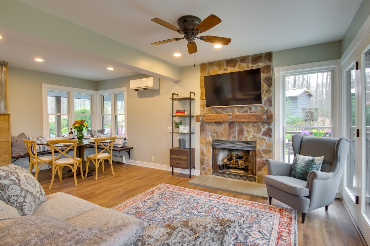 A bright and inviting living area features a comfortable seating arrangement, with a patterned area rug underfoot. A stone fireplace complements the space, while a wall-mounted TV is readily visible. Large windows provide access to natural light, enhancing the space's overall warmth.