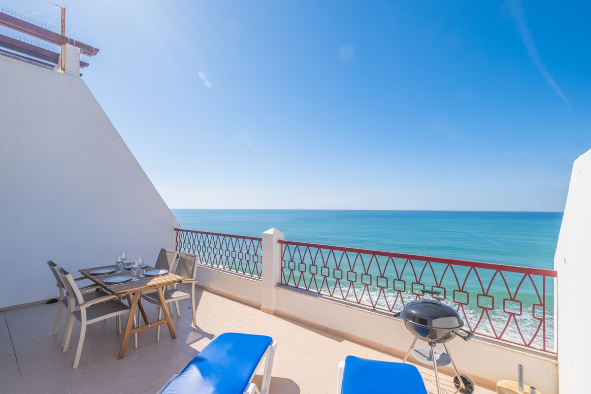 An outdoor terrace offers expansive views of the ocean, featuring a table set for dining and lounging chairs. A barbecue grill is positioned nearby, with a railing providing safety while enjoying the refreshing sea breeze.
