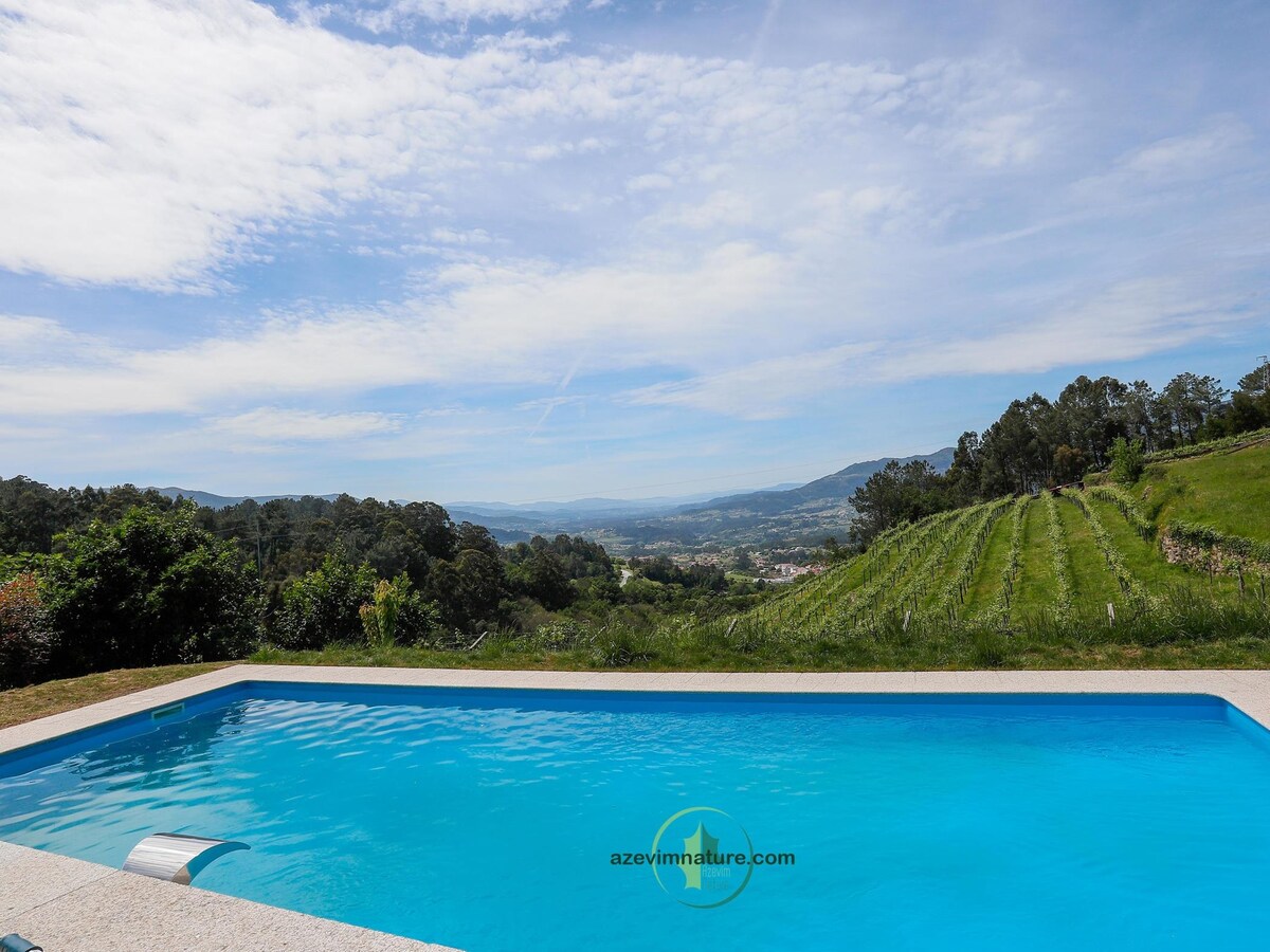A sparkling swimming pool is featured in the foreground, reflecting the sky. Rolling vineyards and lush greenery create a scenic backdrop, with hills and valleys extending into the distance under a clear blue sky.