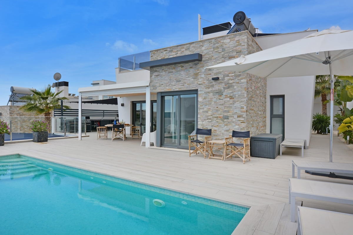 The villa's exterior showcases a modern design with a private swimming pool surrounded by wooden decking. Comfortable seating arrangements near the pool and a dining area are visible under shaded umbrellas. Beautiful landscaping complements the overall aesthetic, providing a relaxing outdoor space.