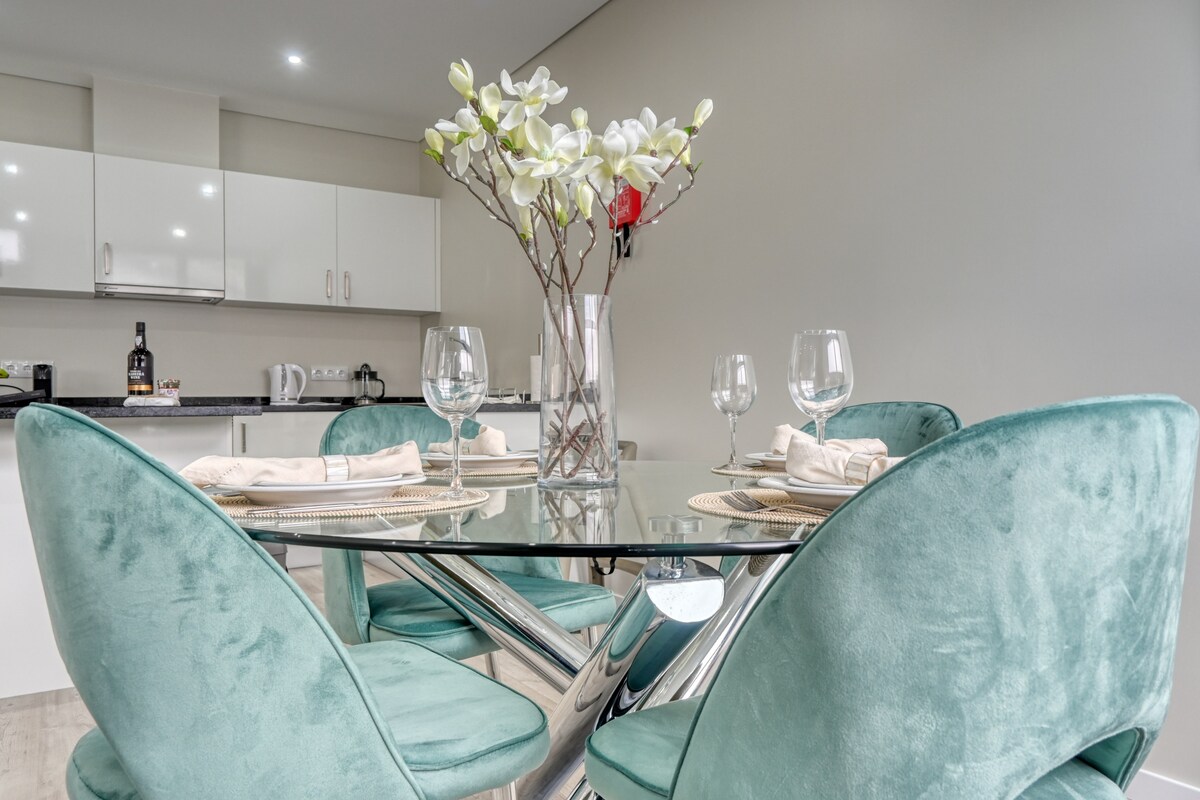A glass dining table is surrounded by four plush, teal chairs. A vase with decorative flowers sits at the center, accompanied by neatly arranged dinnerware and glassware for a welcoming dining experience. The modern kitchen cabinetry is visible in the background.