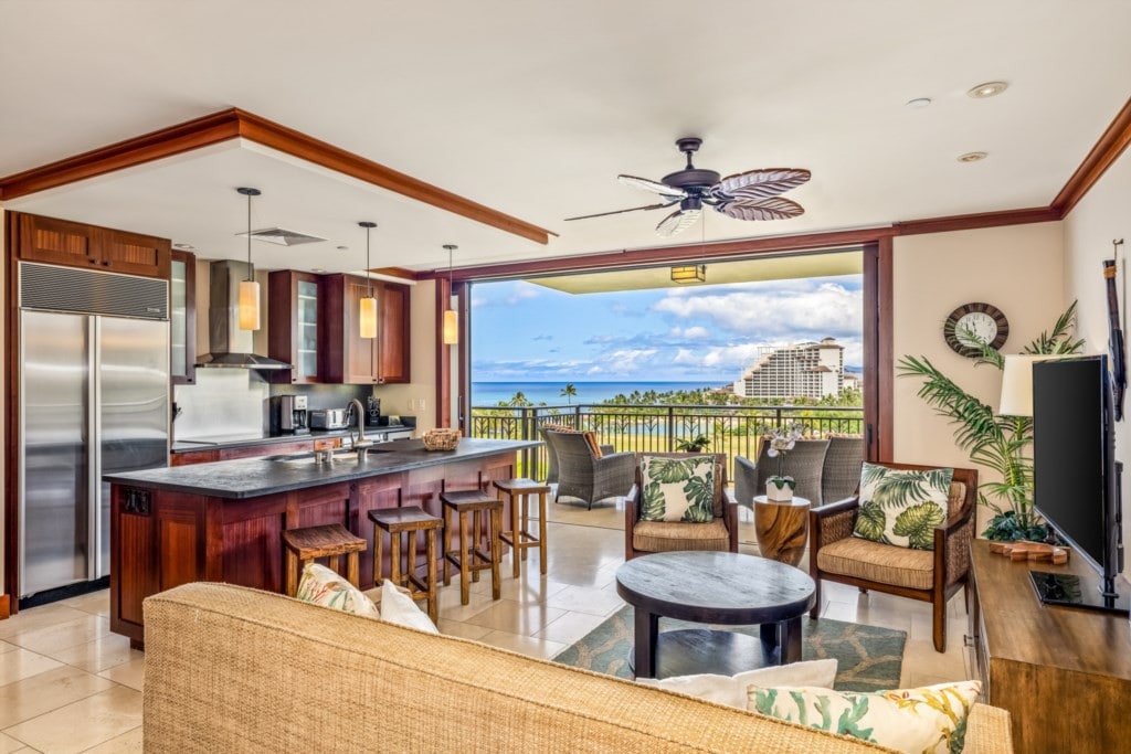 An open-concept living space is shown, featuring a kitchen with modern appliances and a spacious countertop. Comfortable seating is arranged with tropical accent pillows, and large glass doors provide a stunning view of the ocean and surrounding landscape.