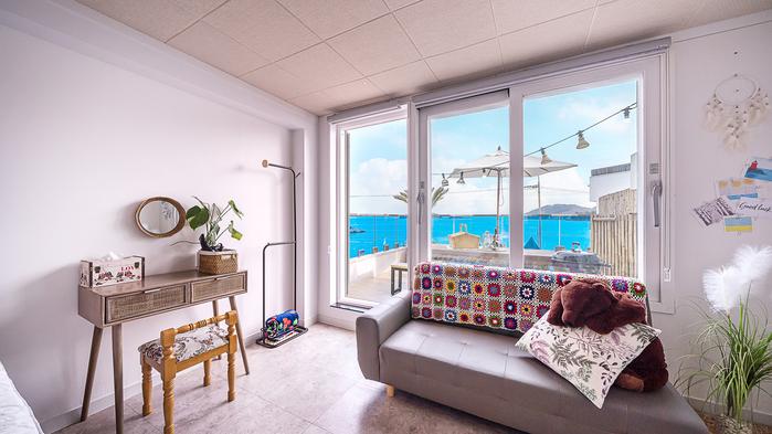 A bright living space features a comfortable sofa adorned with colorful throw pillows. Large glass windows frame a stunning view of the blue sea, while a small table beside a potted plant adds a touch of nature. A cozy atmosphere is created with soft lighting.