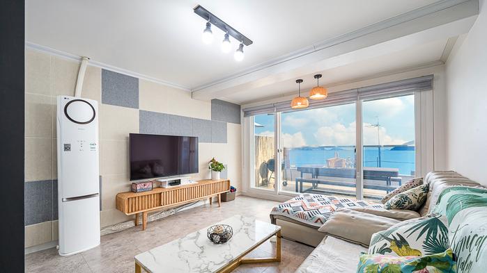 A modern living space is shown, featuring a large window that provides a view of the sea. A flat-screen TV is mounted on the wall. Comfortable seating areas including a sectional sofa and a coffee table are arranged to encourage relaxation and socializing.