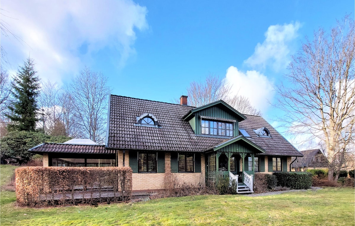 The cottage is set against a blue sky, featuring a charming blend of natural stone and green accents. Large windows and a tiled roof complement the spacious exterior, surrounded by well-maintained grounds and mature trees. A welcoming porch leads to the entrance.