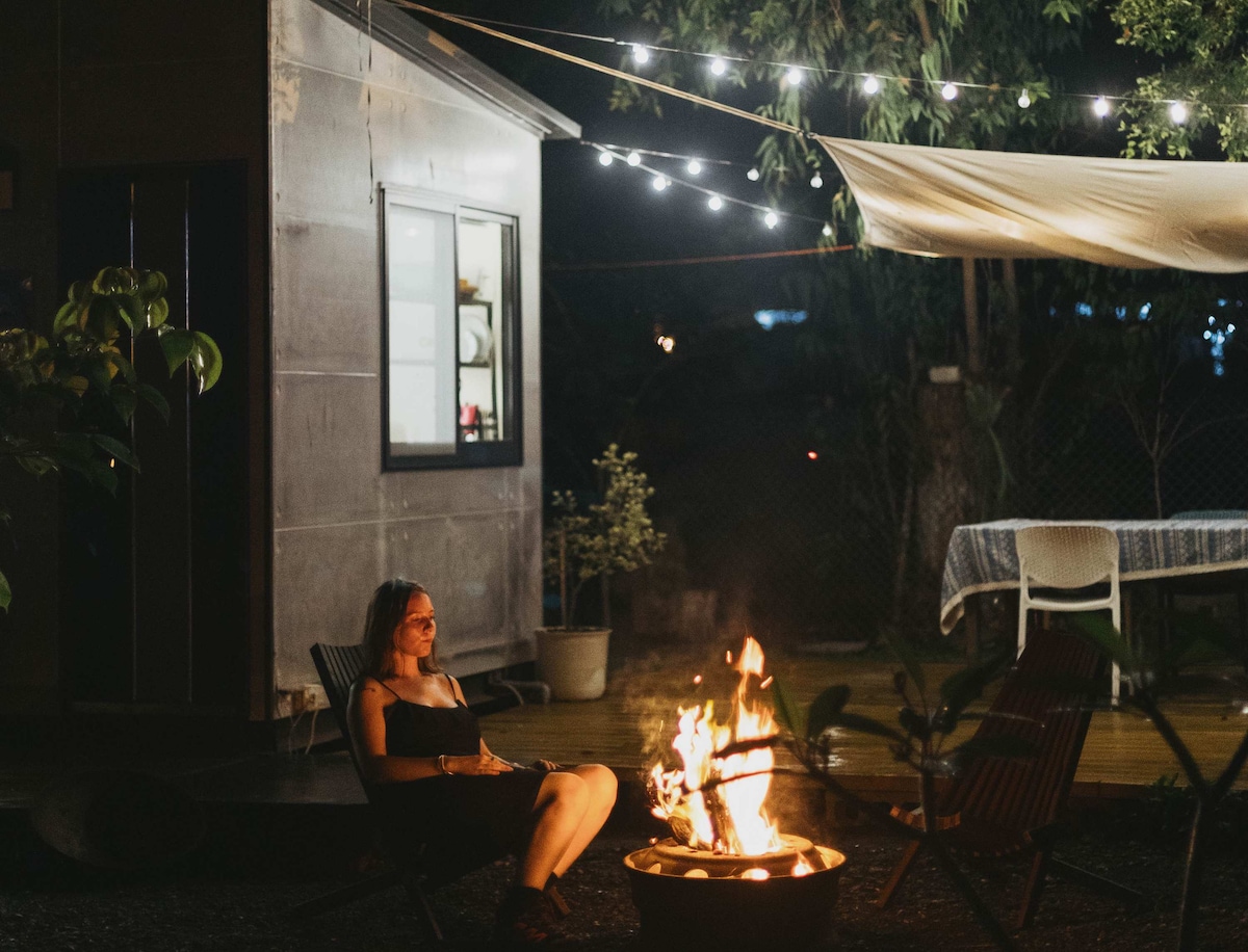 A cozy outdoor setting at night features a fire pit with a warm flame, surrounded by seating on a gravel surface. Soft lights are strung above, illuminating the area, with the cottage visible in the background. A table can be seen nearby, enhancing the inviting atmosphere.