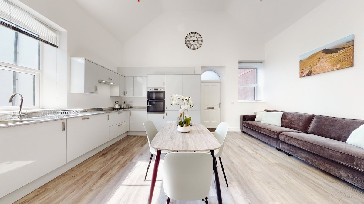 A modern kitchen and dining area showcases an open layout with high ceilings. The light-filled space features a wooden dining table surrounded by chairs. A comfortable sofa is positioned near a window, enhancing the overall spaciousness of the area.