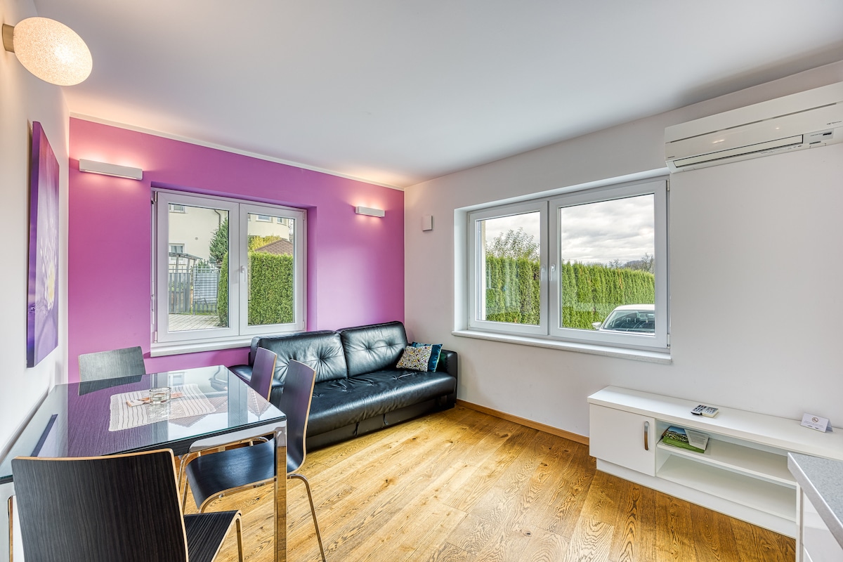 An open-plan living area features a black leather sofa and a glass dining table with contrasting chairs. Two windows allow natural light to fill the space, while a wooden floor adds warmth. Light purple walls create a modern atmosphere.