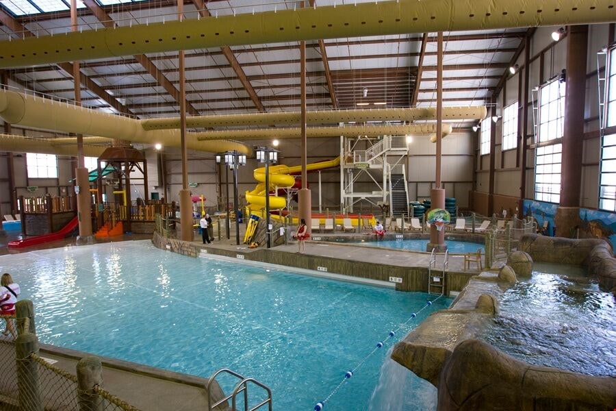 An indoor water park area is visible, featuring a large pool with slides and water features. Brightly colored slides and play structures are located in one corner, while lounge chairs are arranged by the poolside. Natural light streams through high windows, creating a lively atmosphere.