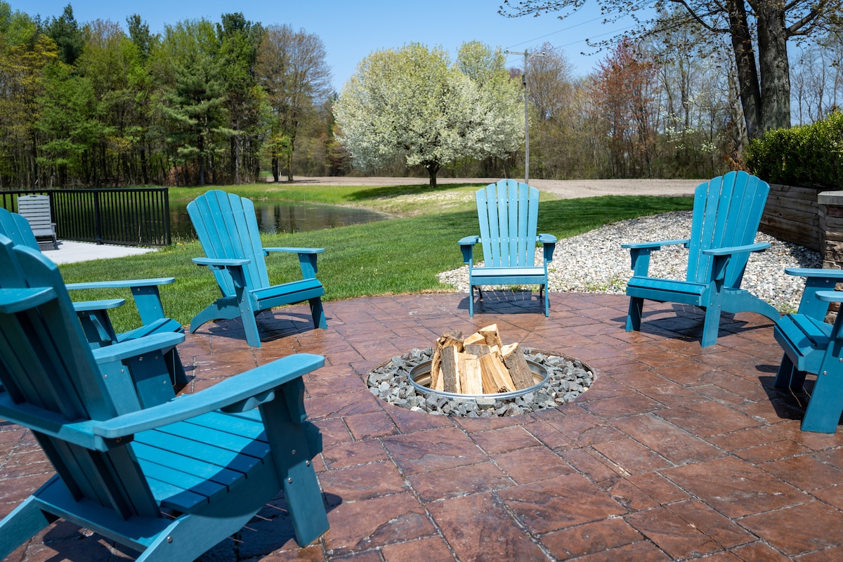 A circular fire pit area features six blue Adirondack chairs arranged around a stone-lined fire pit. The spacious outdoor setting includes a manicured lawn and a serene backdrop of trees and blooming flowers, contributing to a peaceful retreat atmosphere.